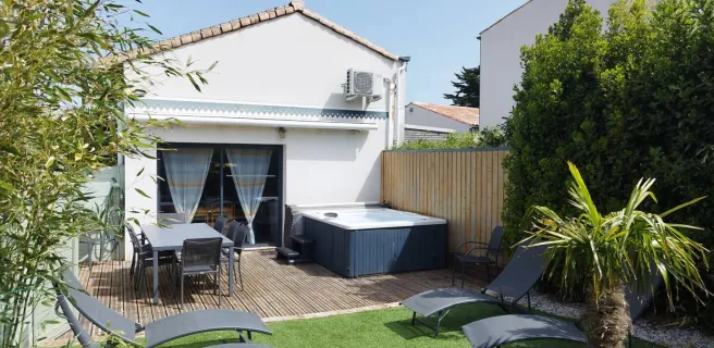 Un jardin avec terrasse en vois et fausse pelouse, avec 4 bains de soleil, une table et 8 chaises, un jaccuzzi. Dans le fond la maison avec porte fenêtre.