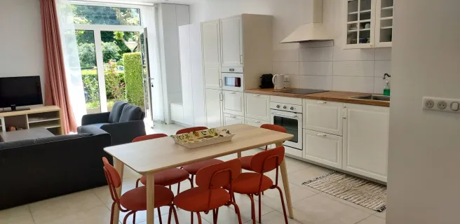 Une pièce de vie ouverte et lumineuse avec une cuisine équipée avec des armoires blanches, un plan de travail et des électroménagers. Un salon attenant est équipé d'un canapé et d'une télévision. Une table en bois avec des chaises rouges est placée entre les deux espaces, créant ainsi un coin repas convivial.
